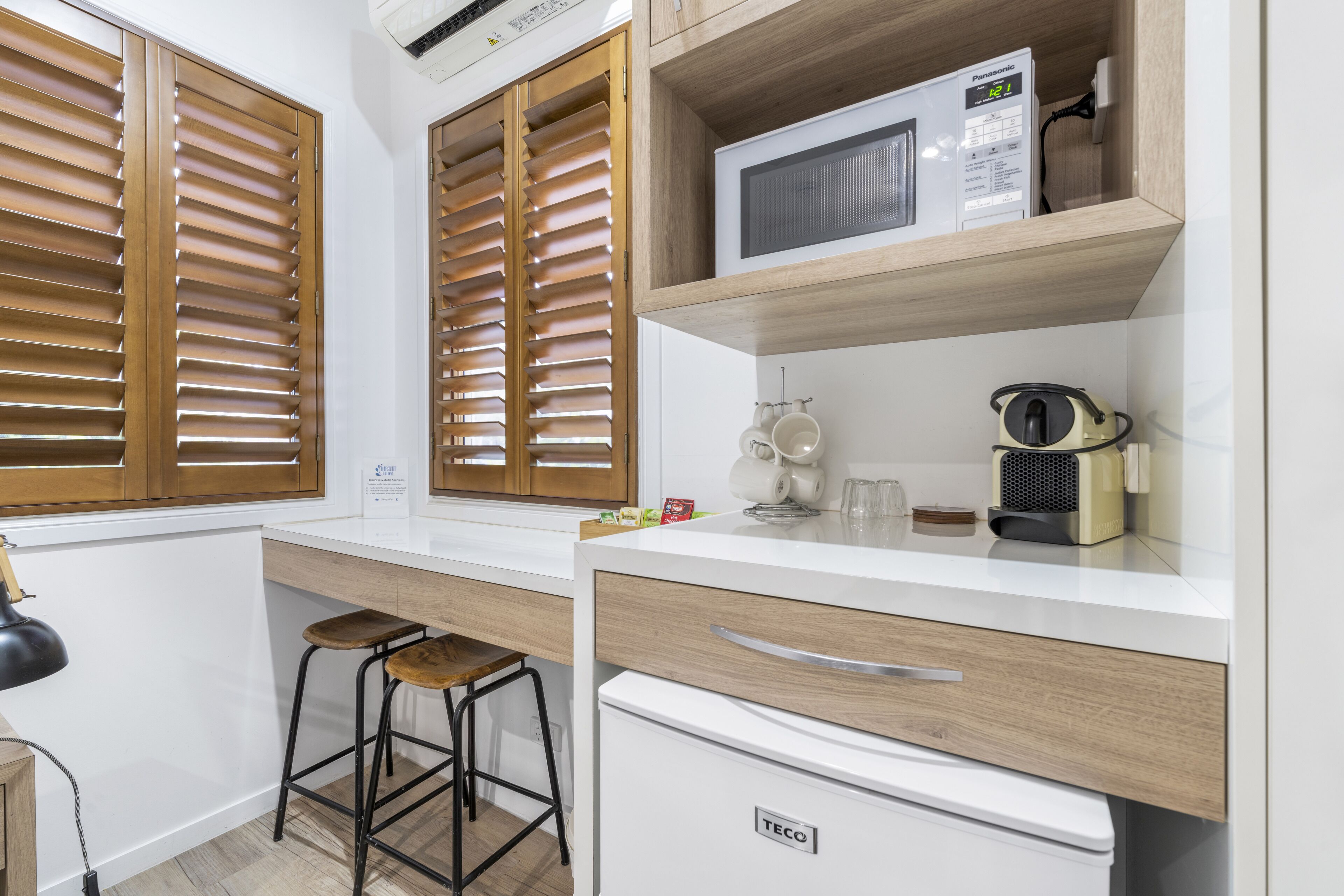 Eine kleine Küche mit Holzjalousien, einem weißen Kühlschrank, einer Mikrowelle und einer Kaffeemaschine.