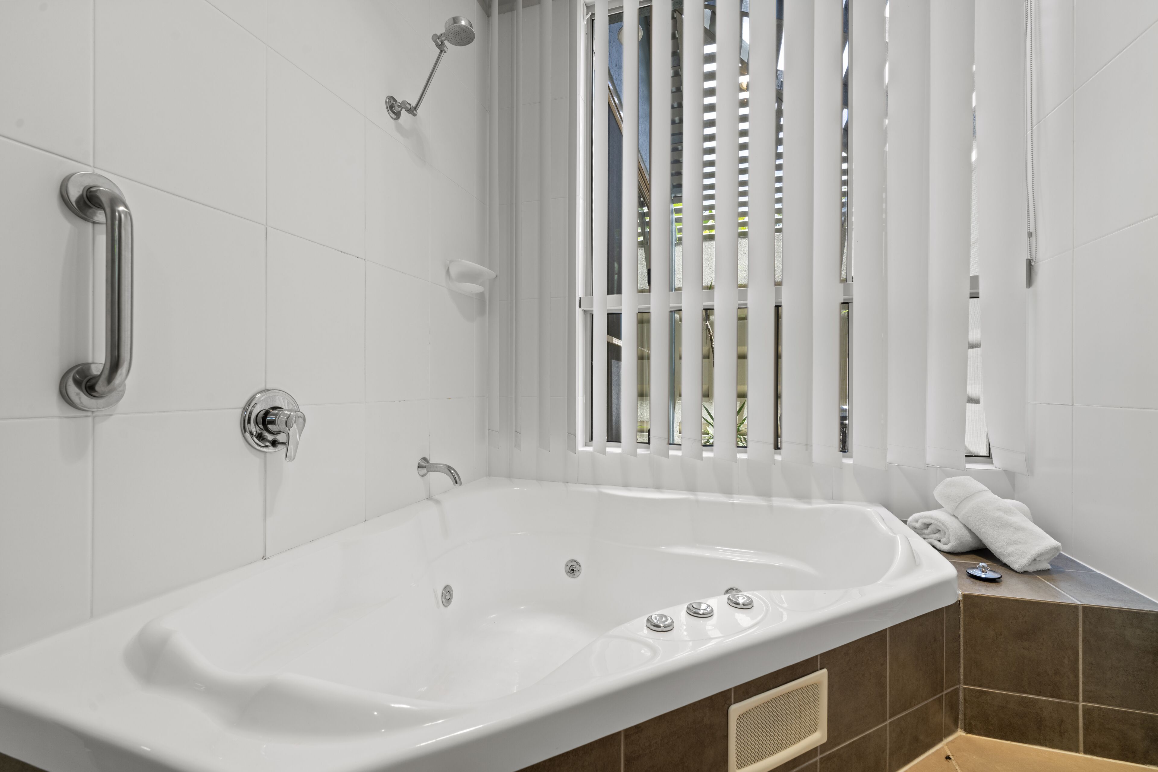 A modern bathroom with a large bathtub, a handheld showerhead, and a window with vertical blinds.