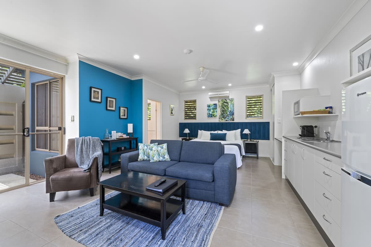 Una sala moderna con sofá, mesa de centro y sillón. Hay una zona de cocina con armarios blancos y un refrigerador. La habitación tiene una pared azul de acento y un ventanal con persianas.