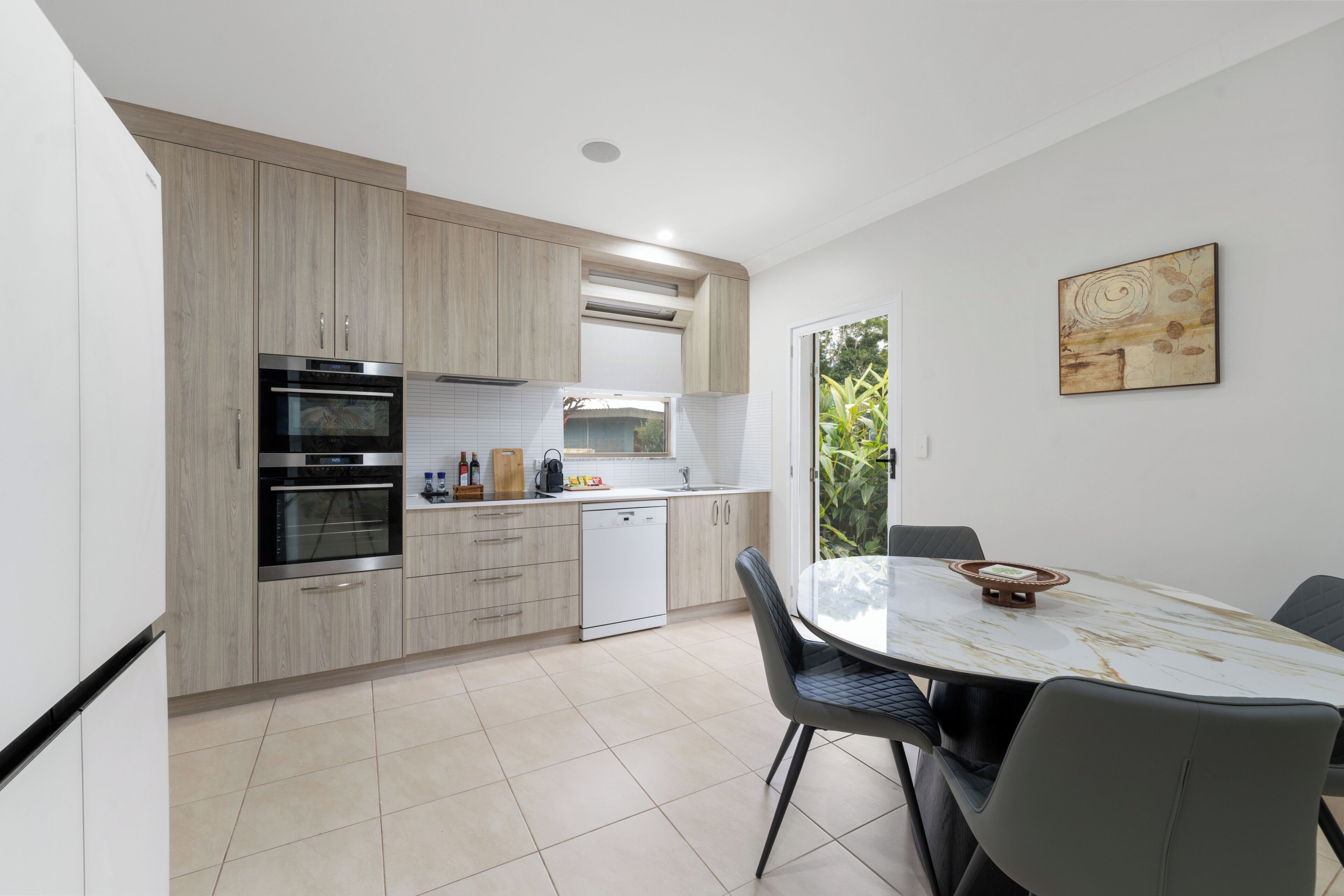 A modern kitchen with built-in appliances, a dining area with a glass table, and a view of greenery outside.