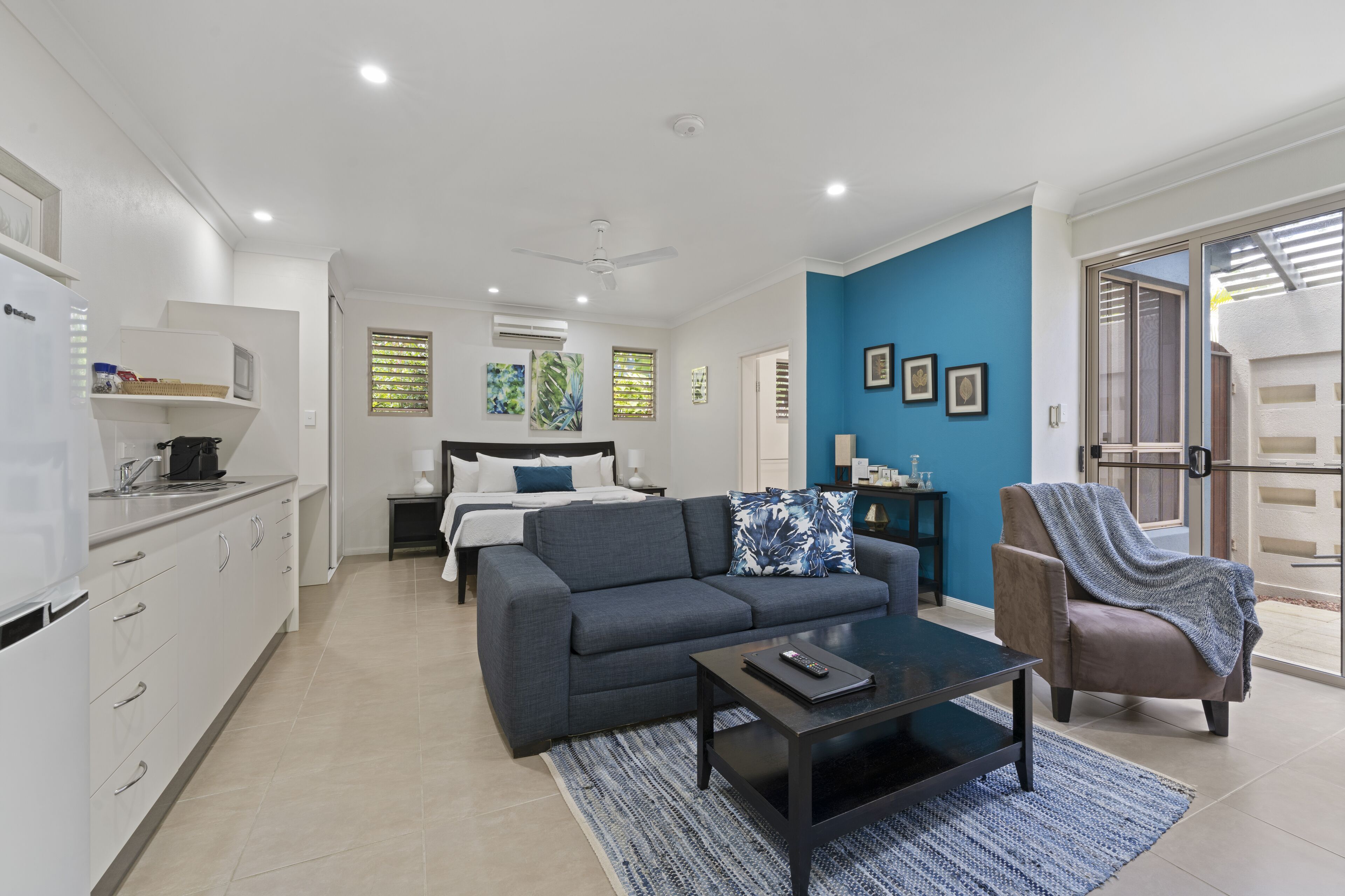 Un salon moderne avec un canapé, une table basse et un fauteuil. Il y a un coin cuisine équipé d’un réfrigérateur et de placards. La pièce est agrémentée d’un mur d’accent bleu et d’une grande fenêtre avec des stores.