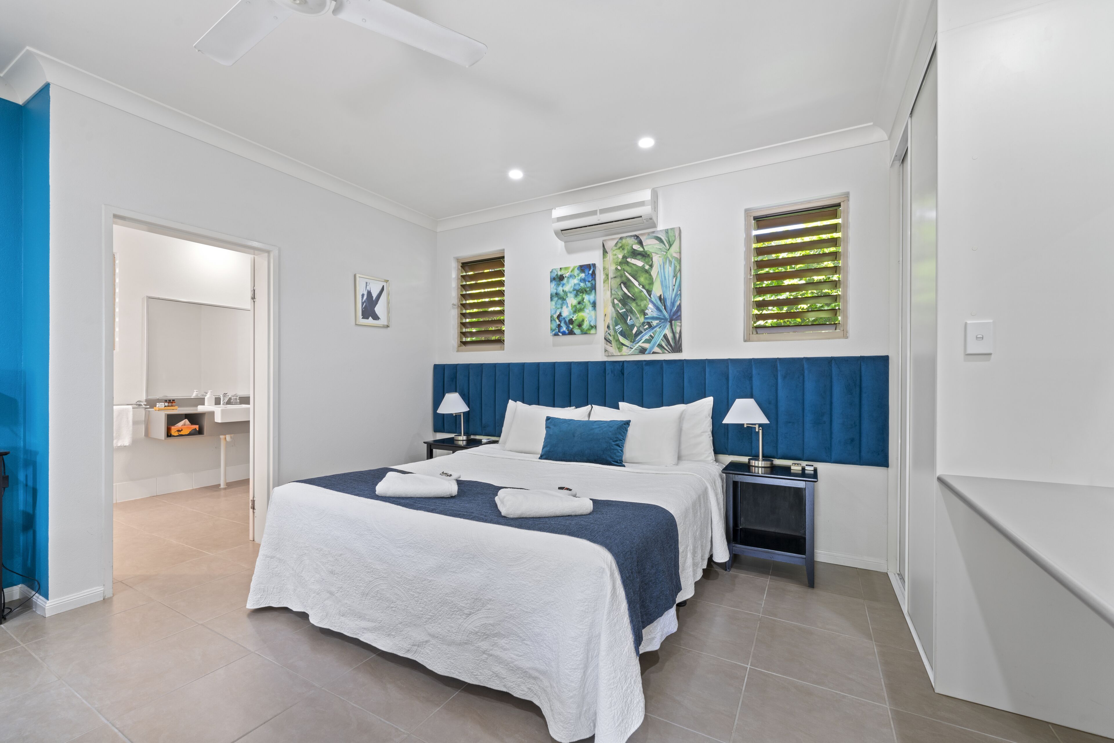 Quarto de hotel moderno com uma cama grande, mesa de cabeceira com abajures, e banheiro visível através de uma porta aberta.