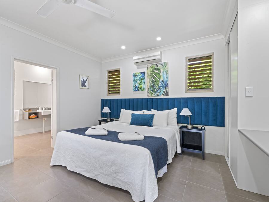 Quarto de hotel moderno com uma cama grande, mesa de cabeceira com abajures, e banheiro visível através de uma porta aberta.