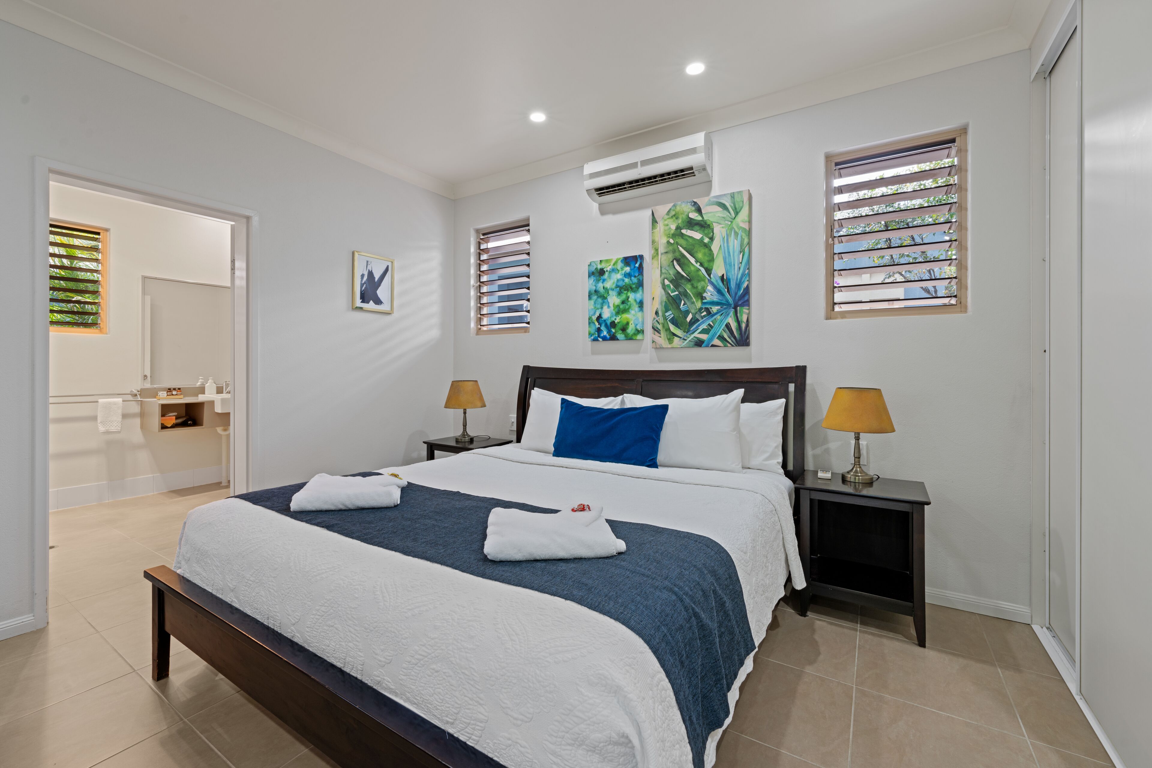 Habitación de hotel moderna con una cama grande, dos mesitas de noche con lámparas, un baño visible a través de una puerta abierta y obras de arte en las paredes.