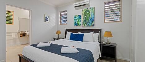 Habitación de hotel moderna con una cama grande, dos mesitas de noche con lámparas, un baño visible a través de una puerta abierta y obras de arte en las paredes.
