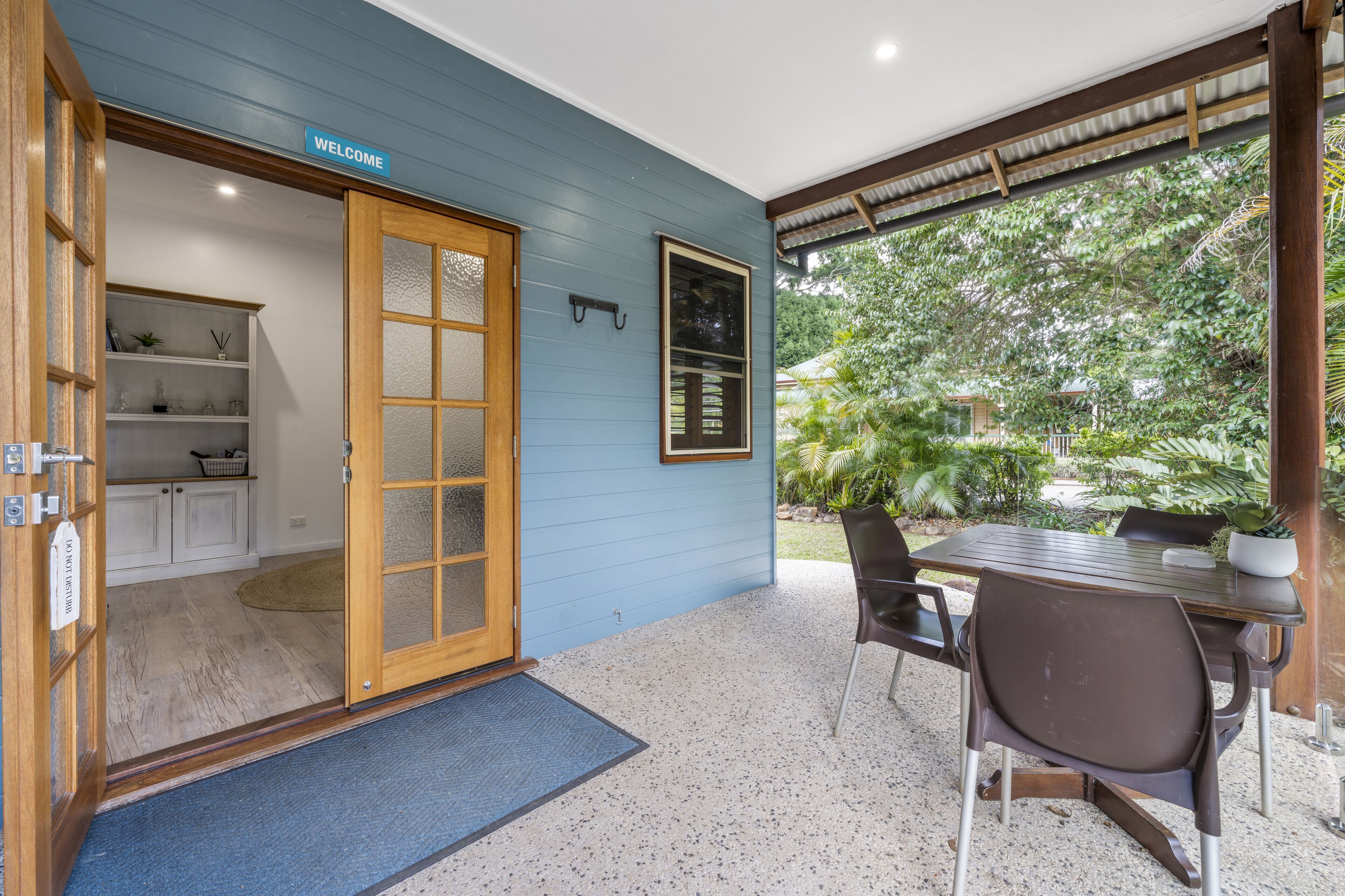 A room with a blue exterior wall, a wooden door, a welcome sign, a kitchen area with white cabinets, and a dining area with a table and chairs.