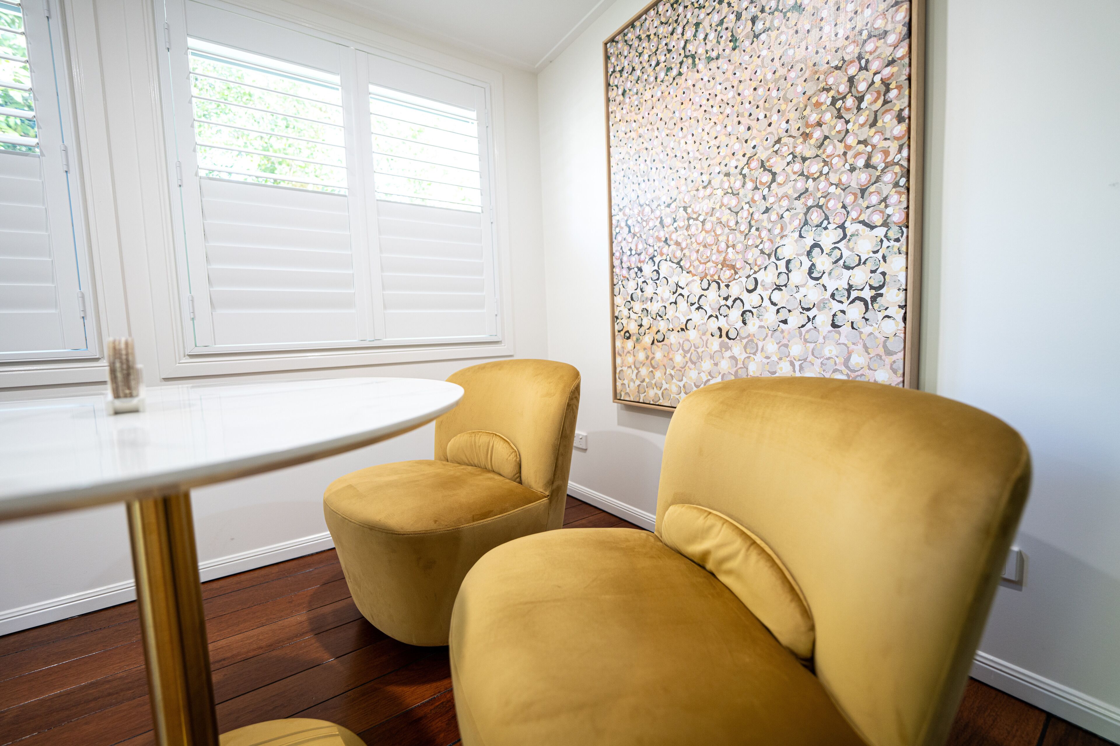 Una habitación con una mesa redonda blanca, dos sillones amarillos, un cuadro abstracto grande en la pared y una ventana con persianas.