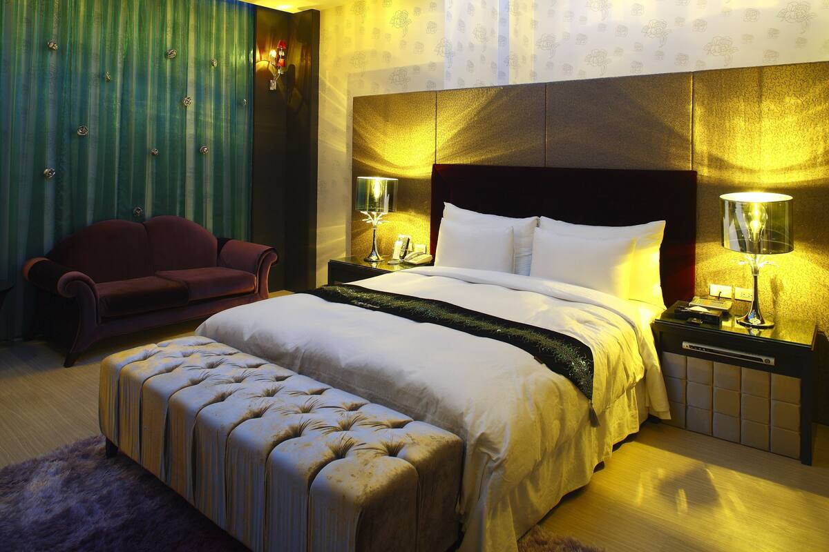 A hotel room with a large bed, a bedside table with lamps, a red sofa, and a patterned wallpaper.