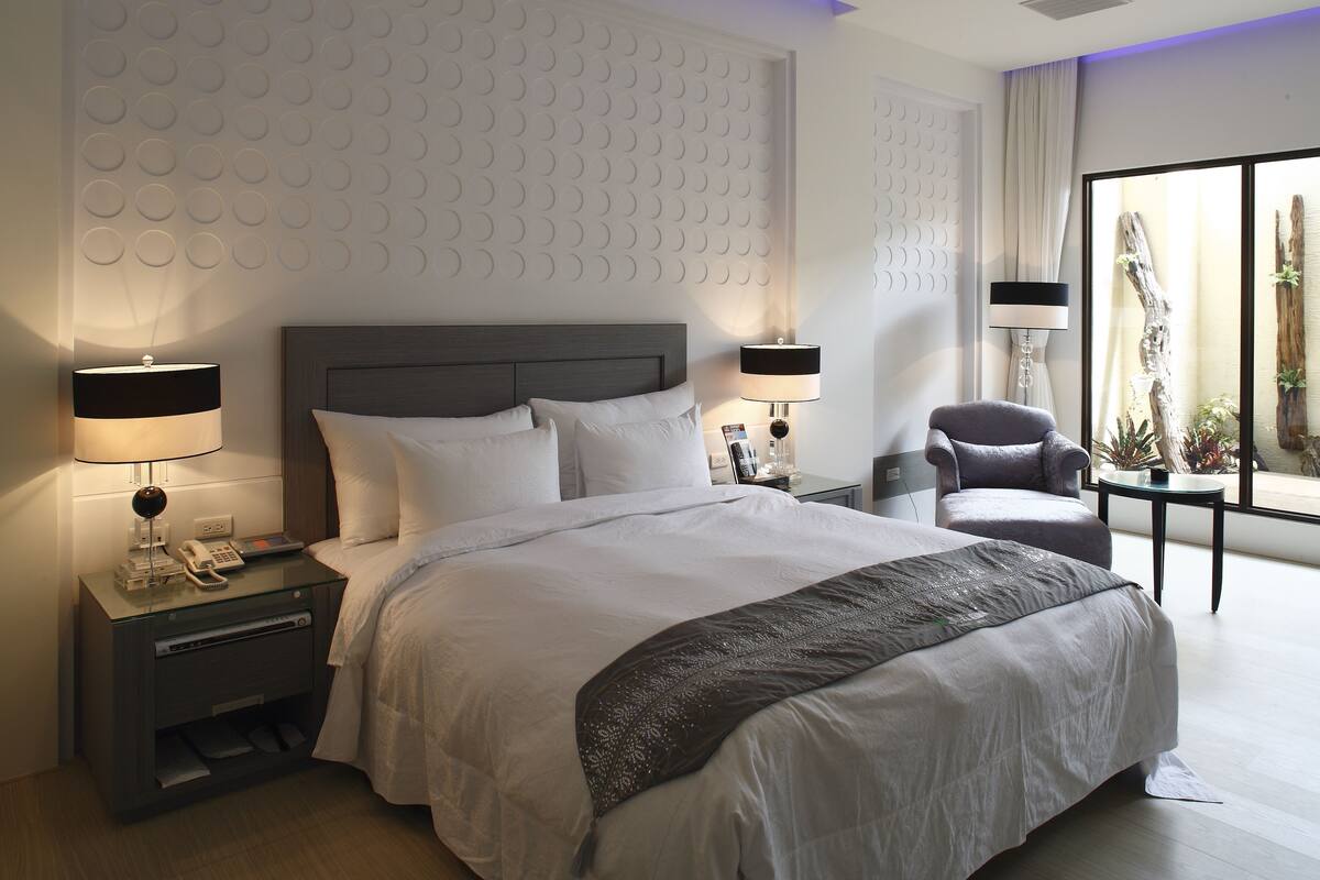A modern hotel room with a large bed, two bedside tables with lamps, a chair, and a large window with a view of the outdoors.