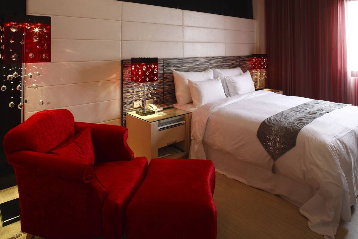 A hotel room with a large bed, a red armchair, a nightstand with a lamp, and a mirror with decorative lights.