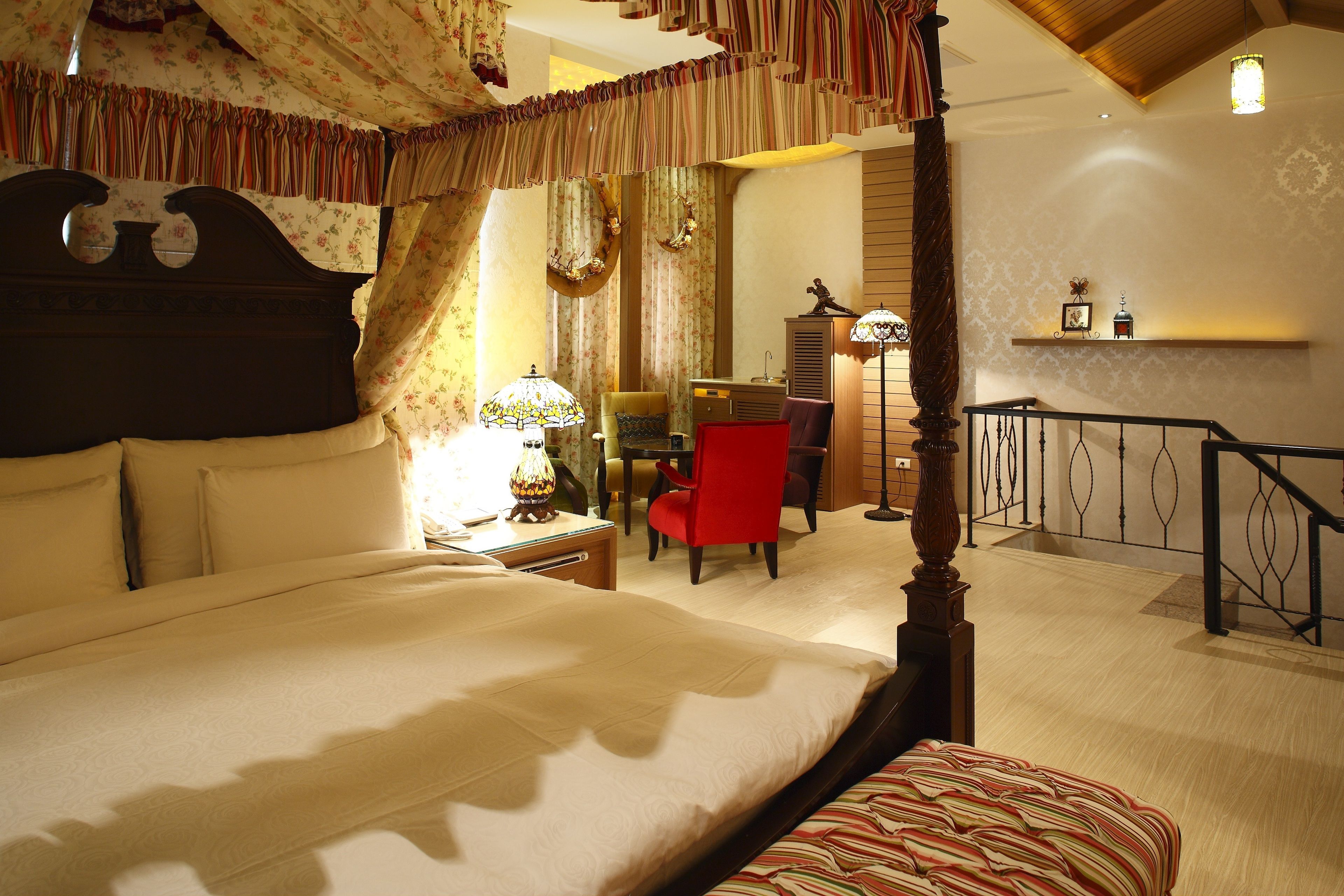 A four-poster bed with a canopy, a desk with a lamp, a red chair, and a small table with a clock.