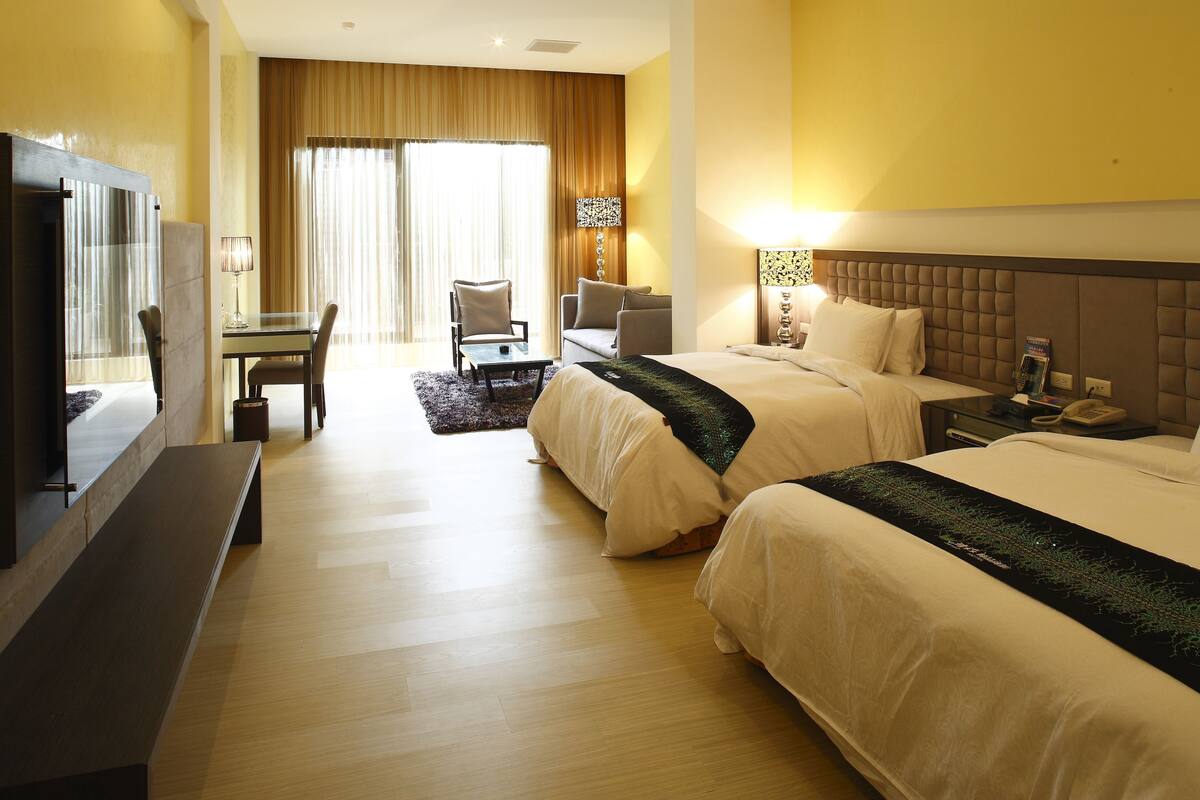 A hotel room with two beds, a desk, a chair, a TV, and a lamp.