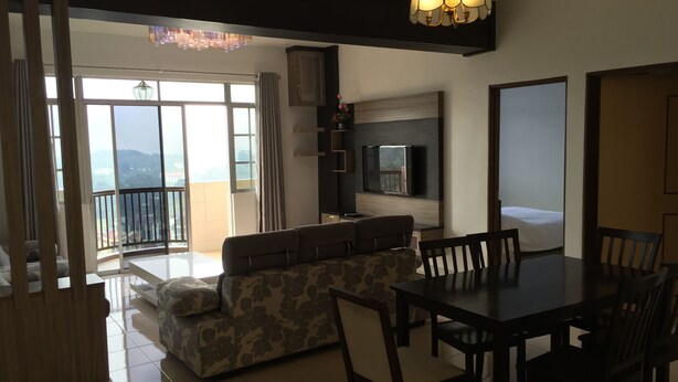 A modern living room with a sofa, dining table, and chairs. There is a TV mounted on the wall, a balcony with a view, and a chandelier. - Cameron Highlands Premier Apartment at Crown Imperial Court (Brinchang)