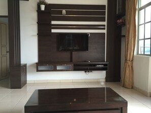 A modern living room with a flat-screen TV mounted on a dark wood panel wall, a glass coffee table in the foreground, and a window with curtains on the right. - Cameron Highlands Premier Apartment at Crown Imperial Court (Brinchang)