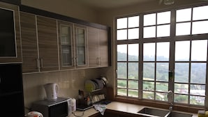 A kitchen with wooden cabinets, a microwave, a kettle, and a window with a view of greenery. - Cameron Highlands Premier Apartment at Crown Imperial Court (Brinchang)