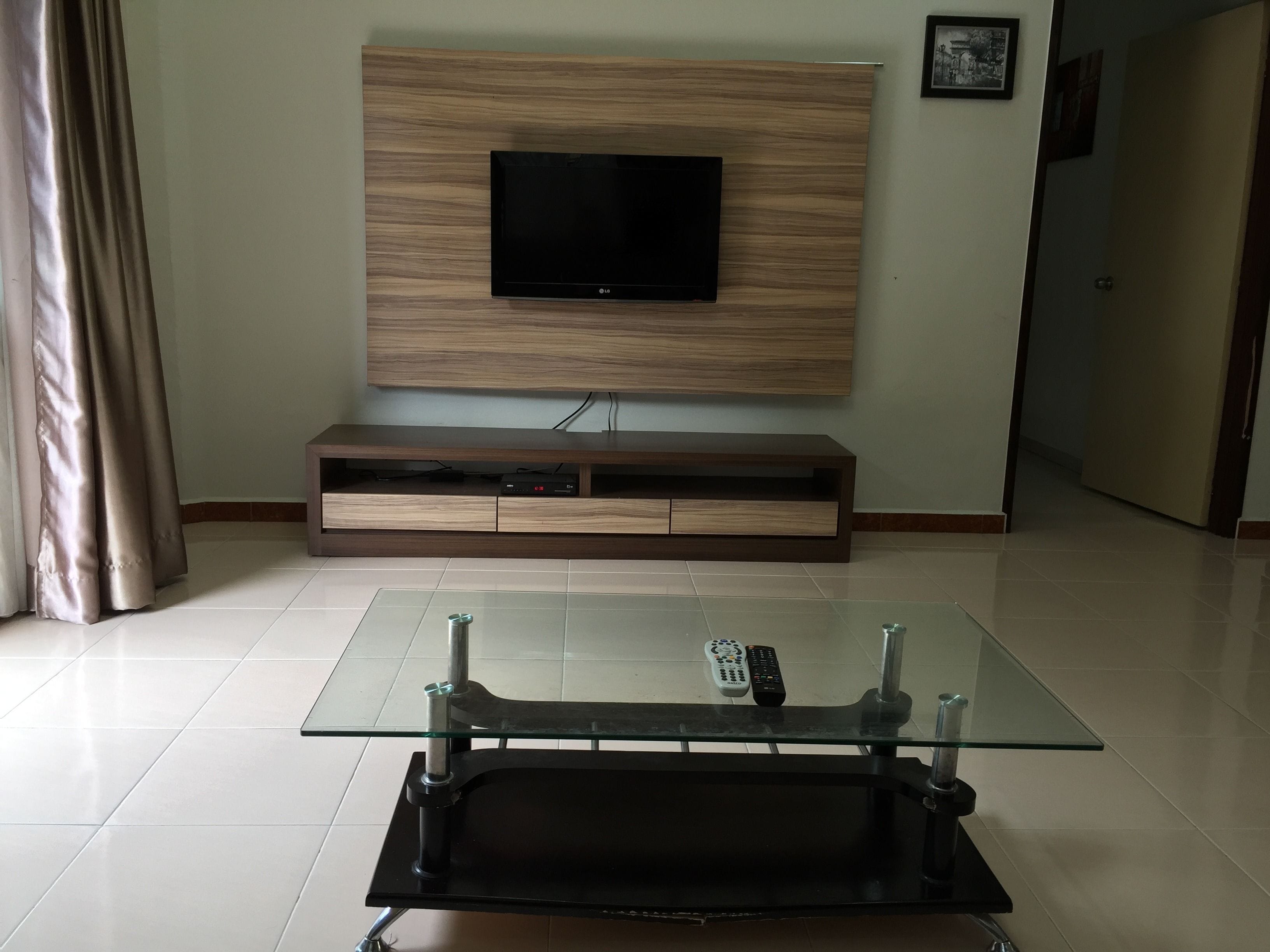 A living room with a flat-screen TV mounted on a wooden wall, a glass coffee table with a remote control, and a door in the background.