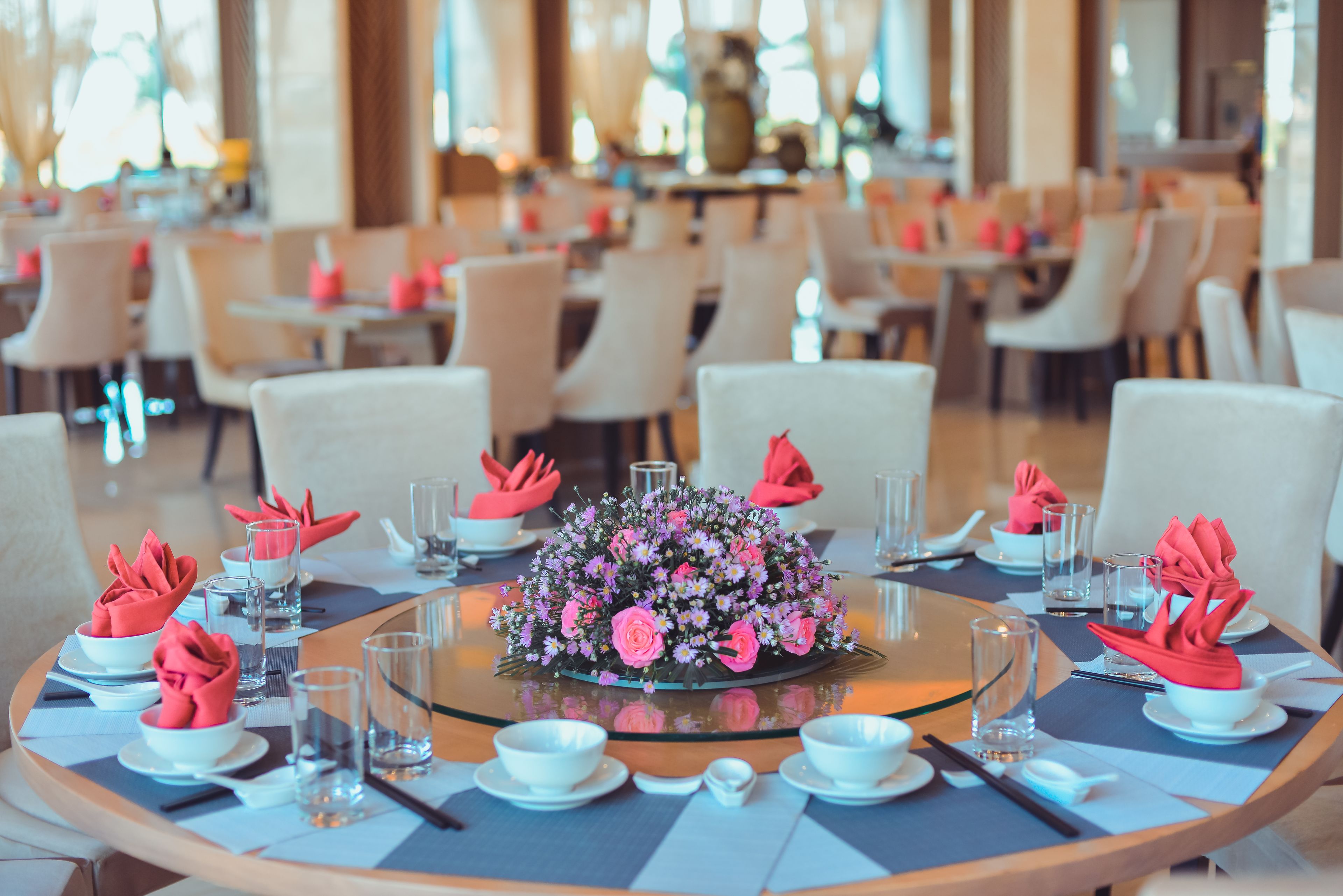 A dining area with round tables set for a meal, each with a centerpiece of pink flowers and red napkins.