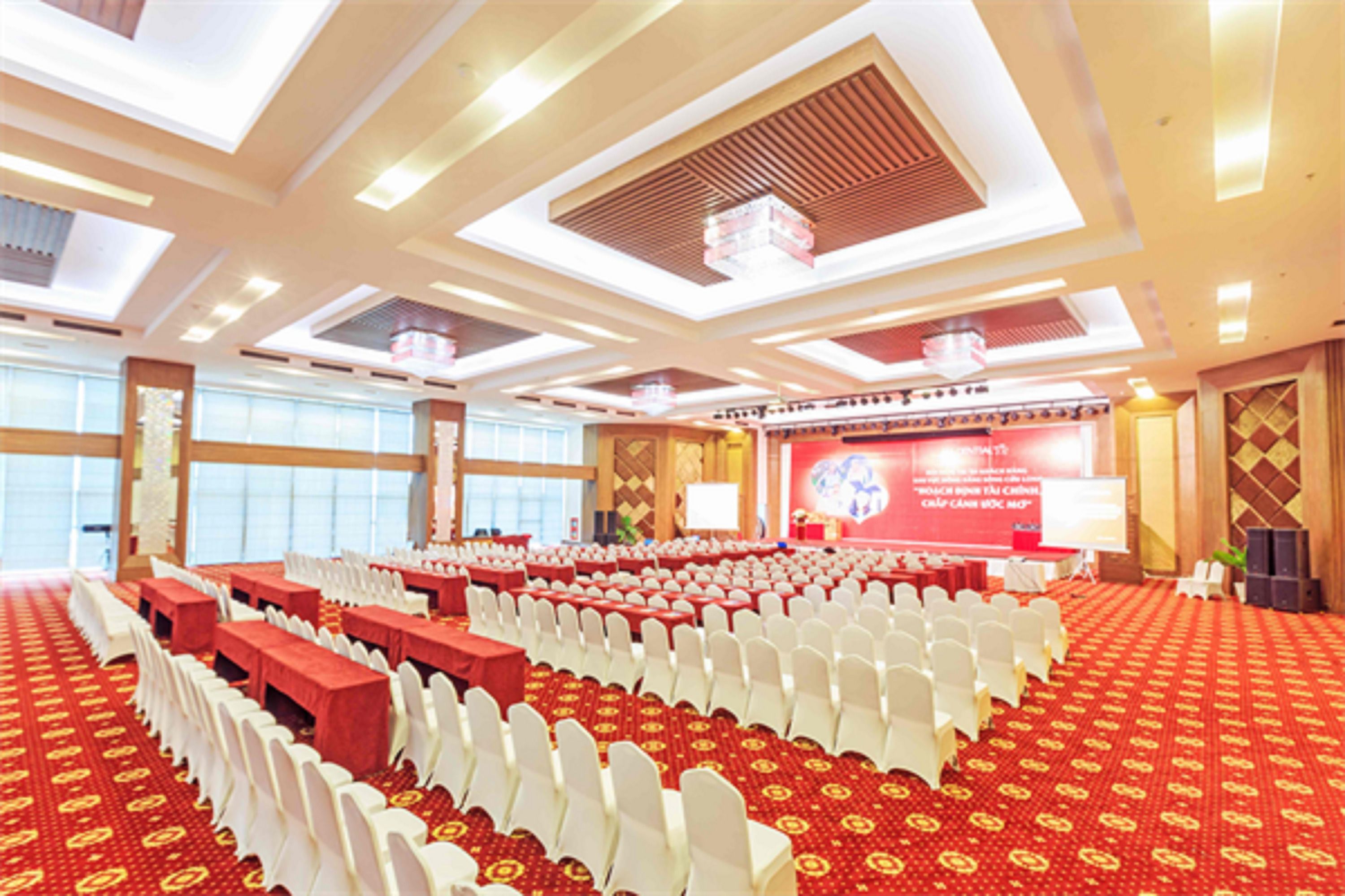 A large conference room with rows of tables and chairs, a stage with a presentation screen, and a patterned carpet.