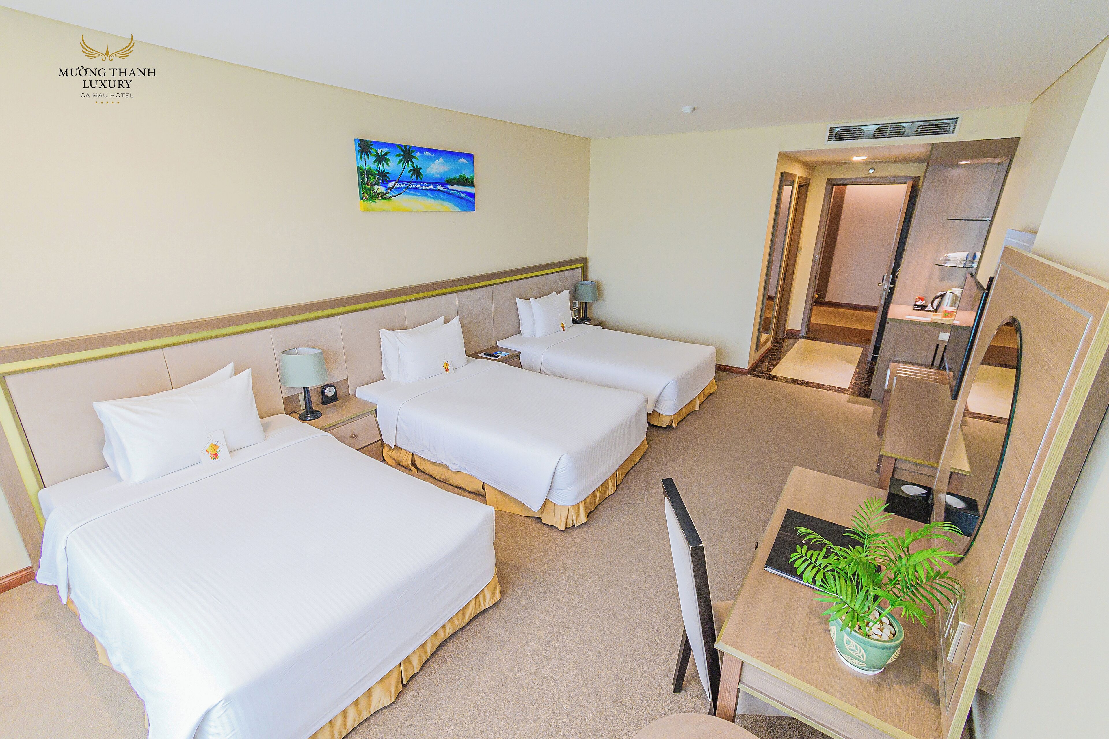 A hotel room with three beds, a desk, a chair, a plant, and a painting on the wall.