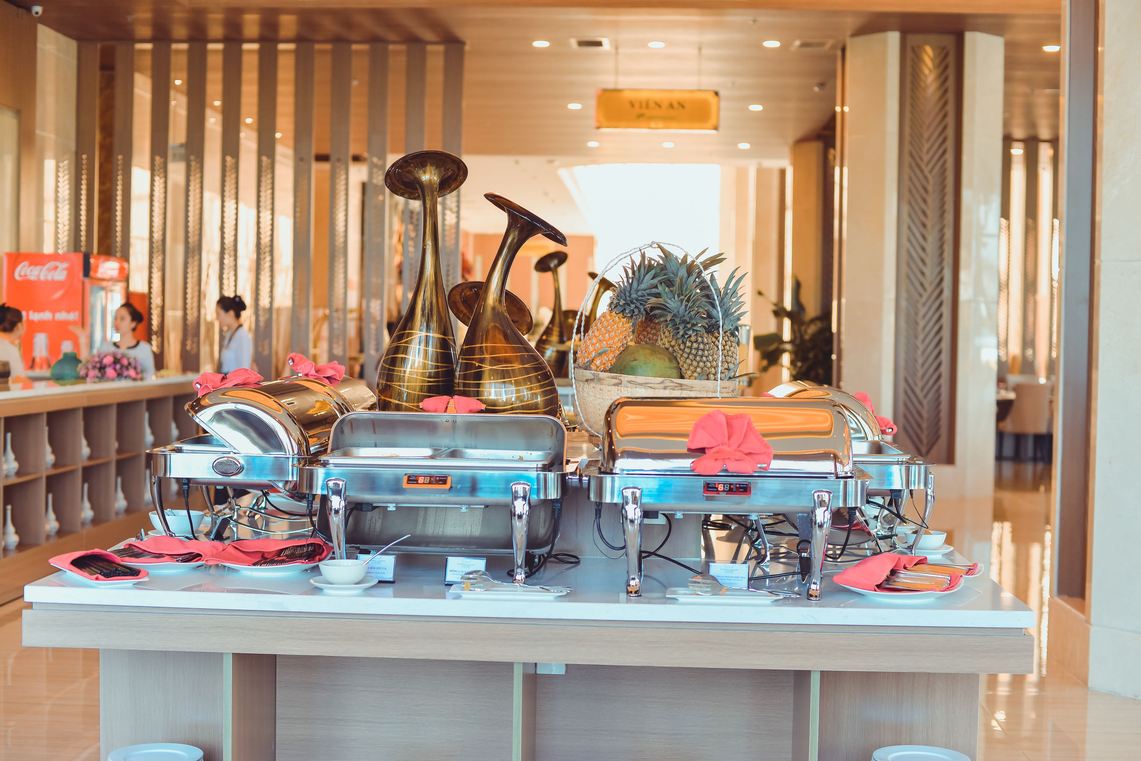 A buffet setup with various food containers and utensils, a fruit basket, and a Coca-Cola refrigerator.