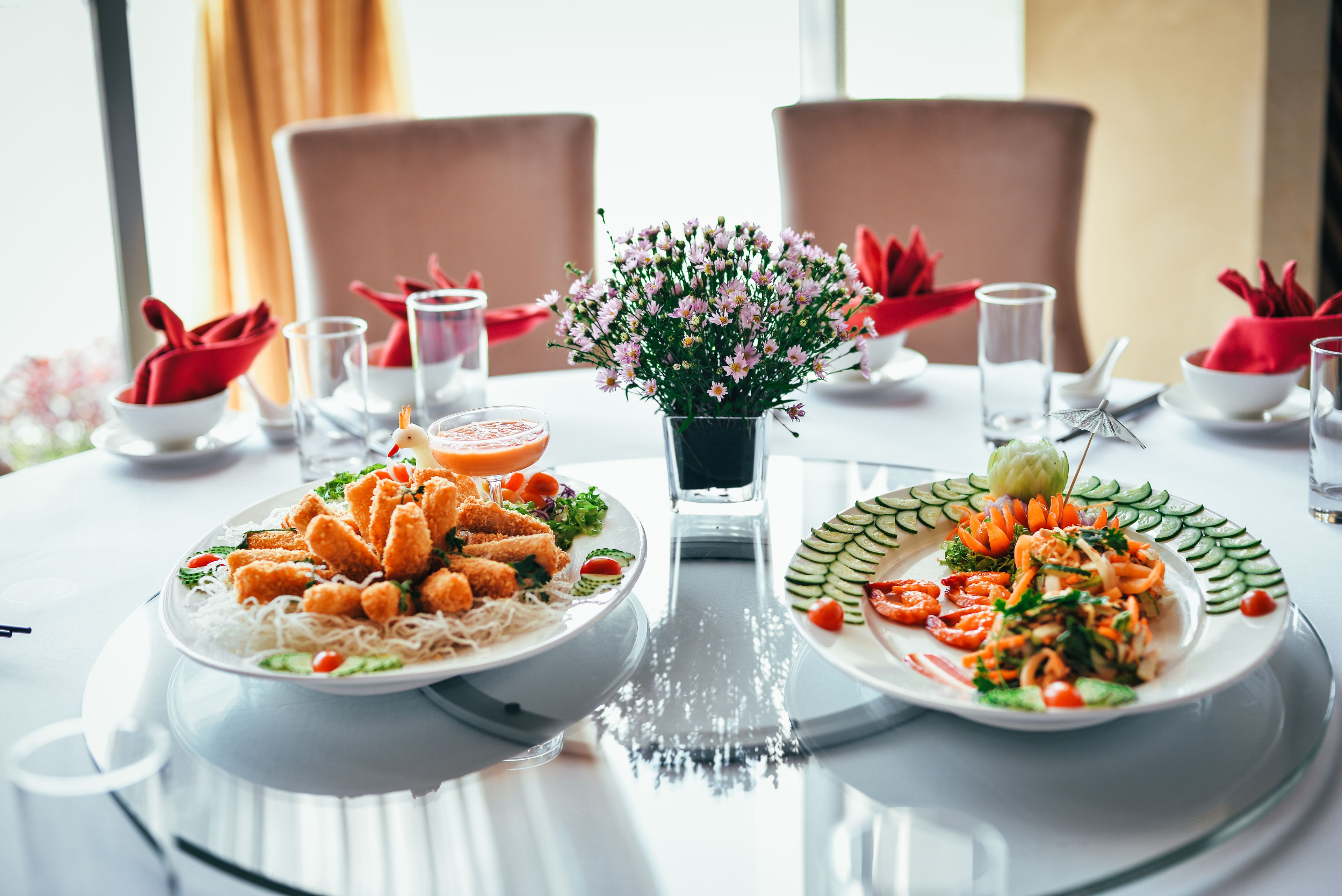 A dining table with two plates of food, a vase of flowers, and red napkins.