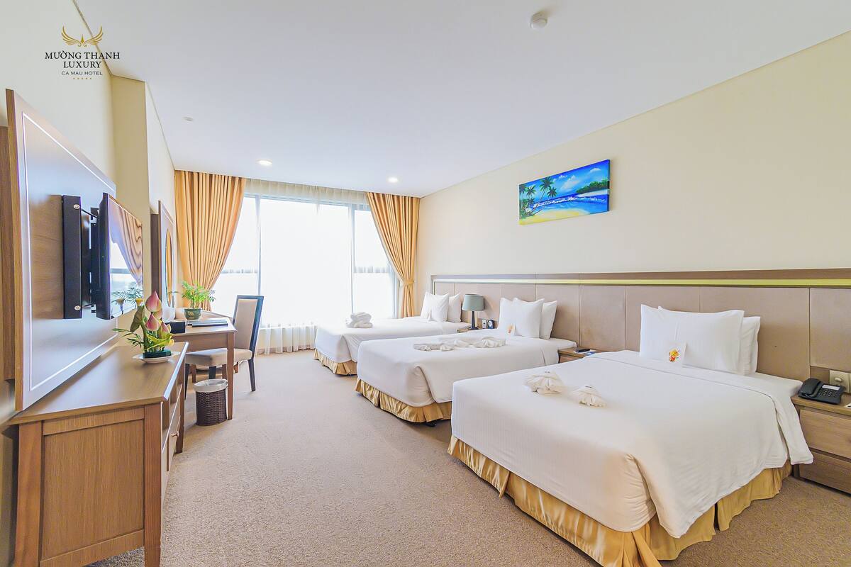 A hotel room with three beds, a desk, a chair, a TV, and a painting on the wall.