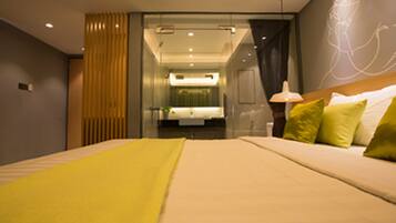 Habitación de hotel moderna con una cama grande, una mesita de noche, una lámpara y un baño visible a través de un tabique de vidrio.