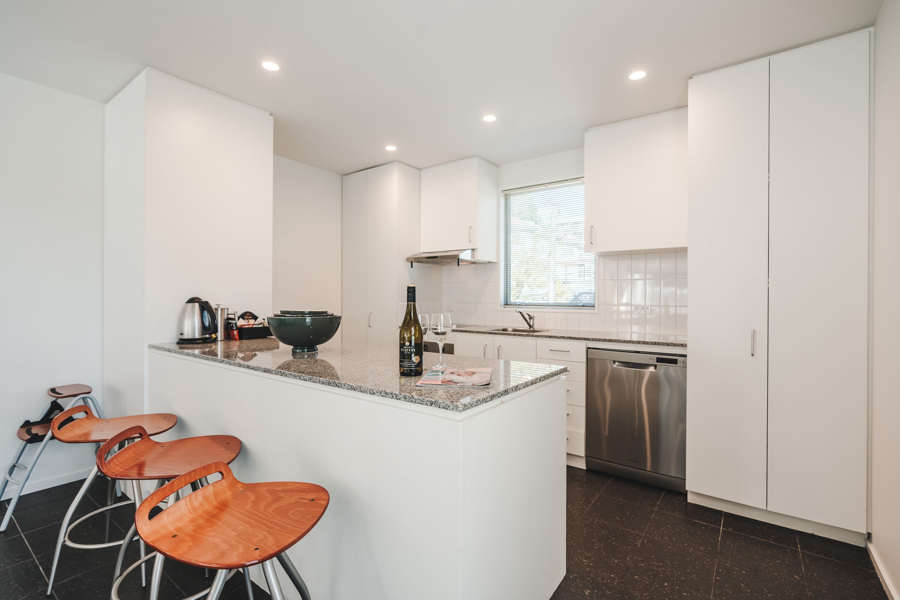 Una cocina moderna con armarios blancos, encimera de granito y lavavajillas integrado. Hay tres taburetes de madera y una botella de vino en la encimera.