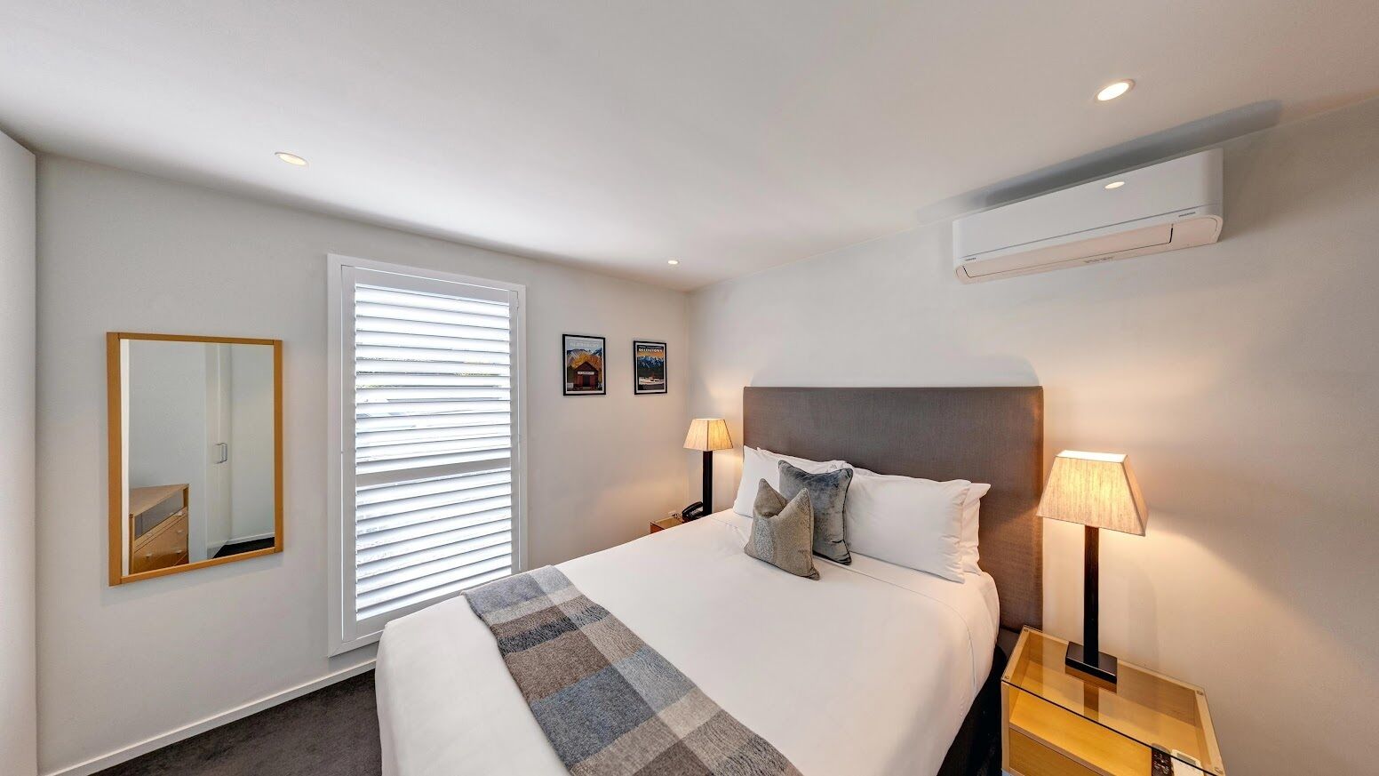 Habitación de hotel con cama, lámparas de noche, espejo, ventana con persianas y aire acondicionado.