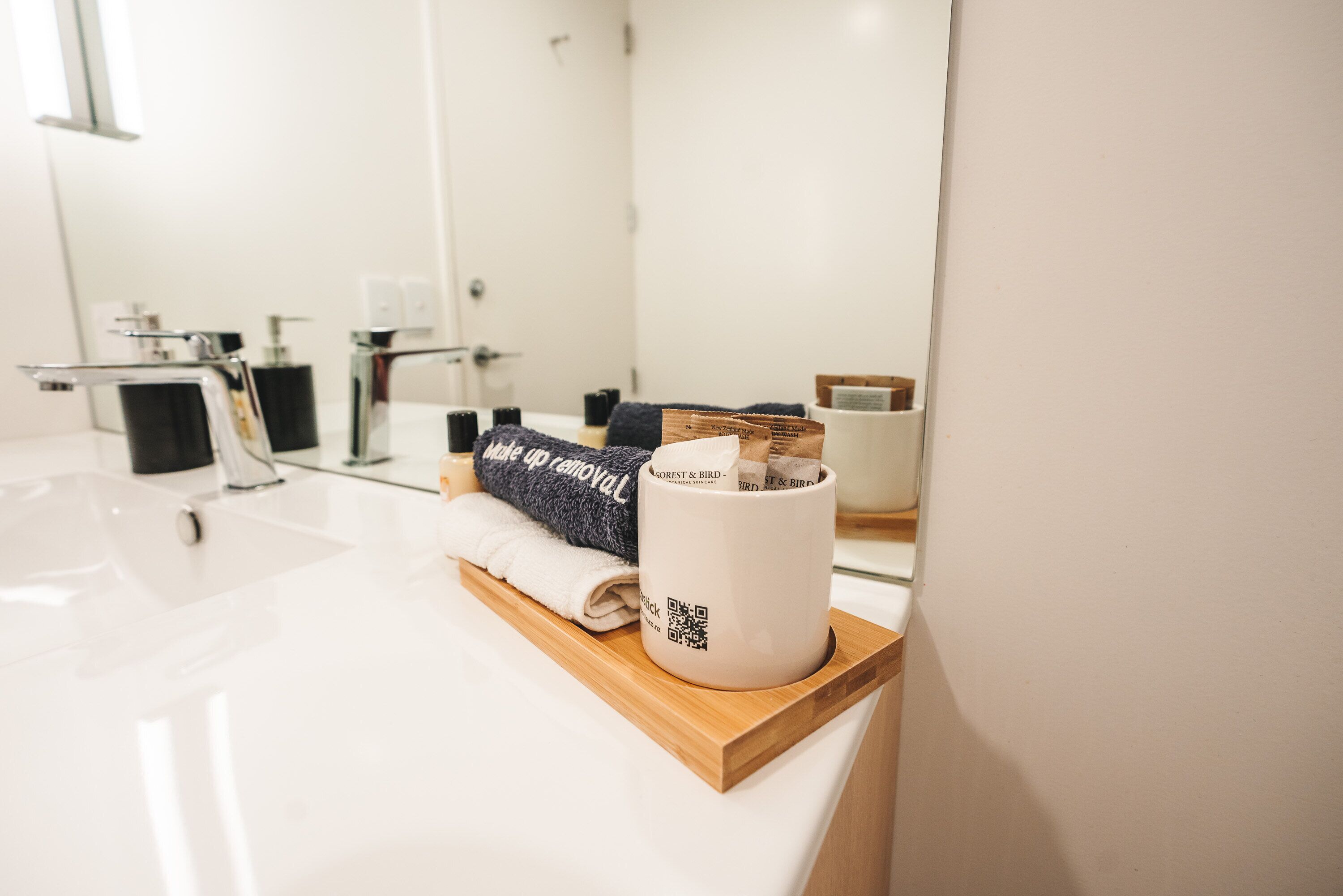 A modern bathroom with a white sink, a wooden tray, a rolled towel, and a mug with a QR code.