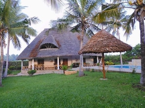 A thatched-roof house with a pool, surrounded by palm trees. - Queen K Cottages (Watamu)