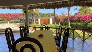 A dining area with a table set for six, surrounded by chairs, with a view of a pool and garden. - Queen K Cottages (Watamu)