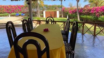 An outdoor dining area with a table set for four, surrounded by chairs, with a view of a swimming pool and lush greenery.