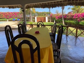 A dining area with a table set for six, surrounded by chairs, with a view of a pool and garden. - Queen K Cottages (Watamu)