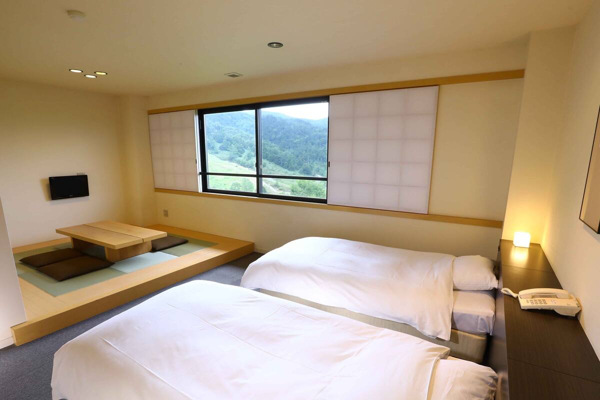 Quarto de hotel com duas camas, um espaço de estar pequeno, janela com vista para as montanhas e telefone ao lado da cama.