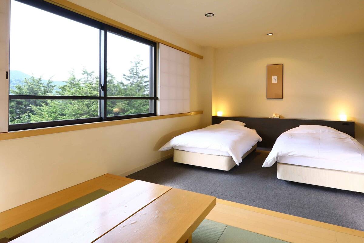 Quarto de hotel com duas camas, janela com vista para vegetação, mesa de cabeceira e luminária fixada na parede.