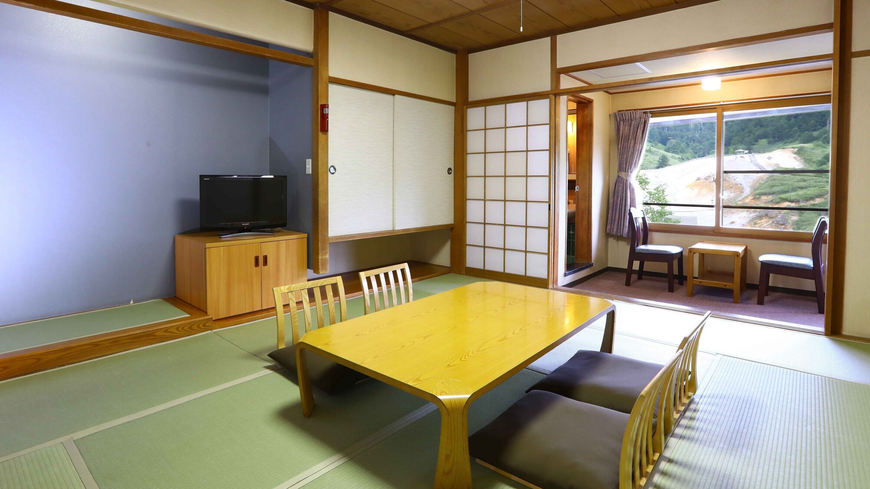 A traditional Japanese room with tatami flooring, a wooden table, chairs, a television, and a view of a landscape through a window.