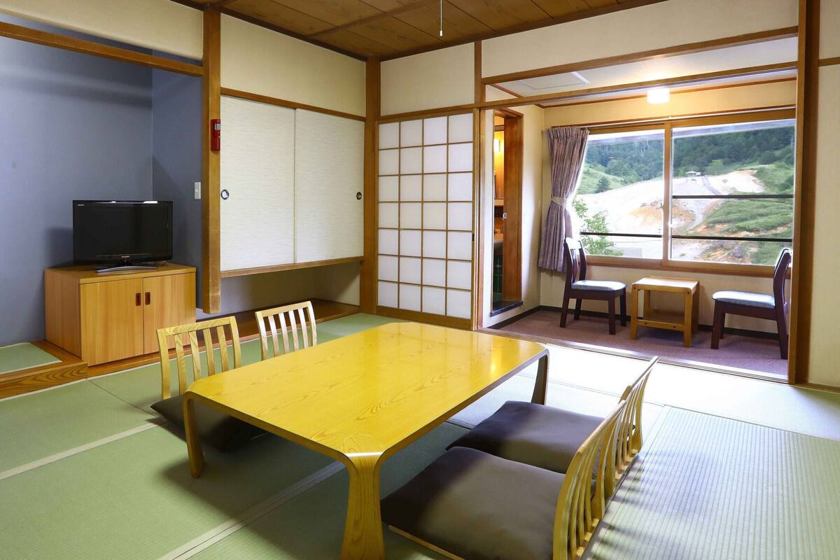 Um quarto japonês tradicional com tatames, uma mesa de madeira, cadeiras, uma televisão e vista para uma paisagem pela janela.