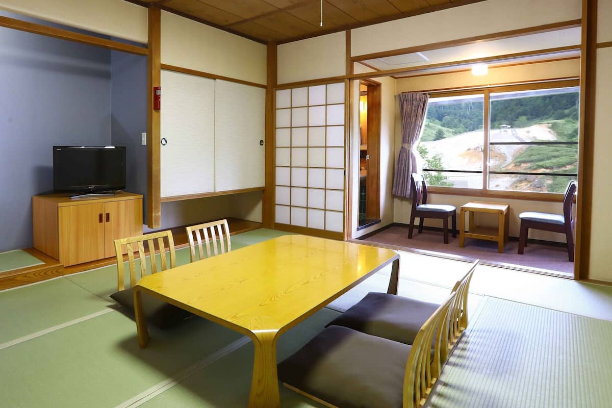 A traditional Japanese room with tatami flooring, a wooden table, chairs, a television, and a view of a landscape through a window.