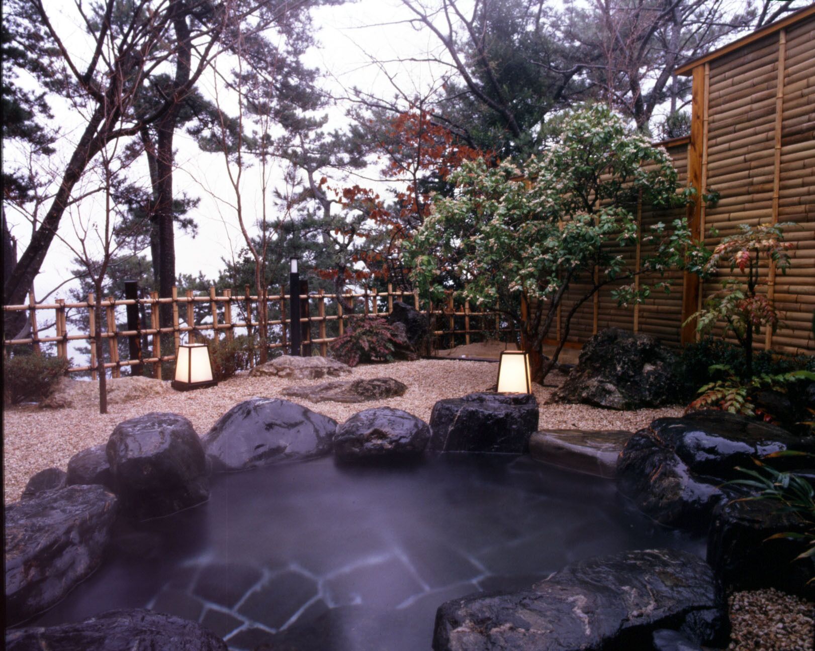 A hot spring surrounded by rocks and trees, with a wooden fence and a building in the background.