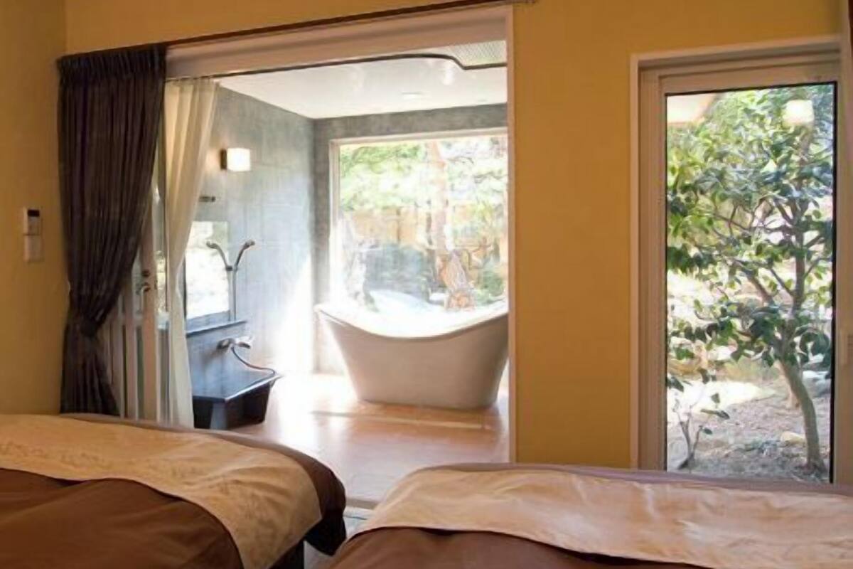 A hotel room with two beds, a bathtub, and a view of greenery outside.