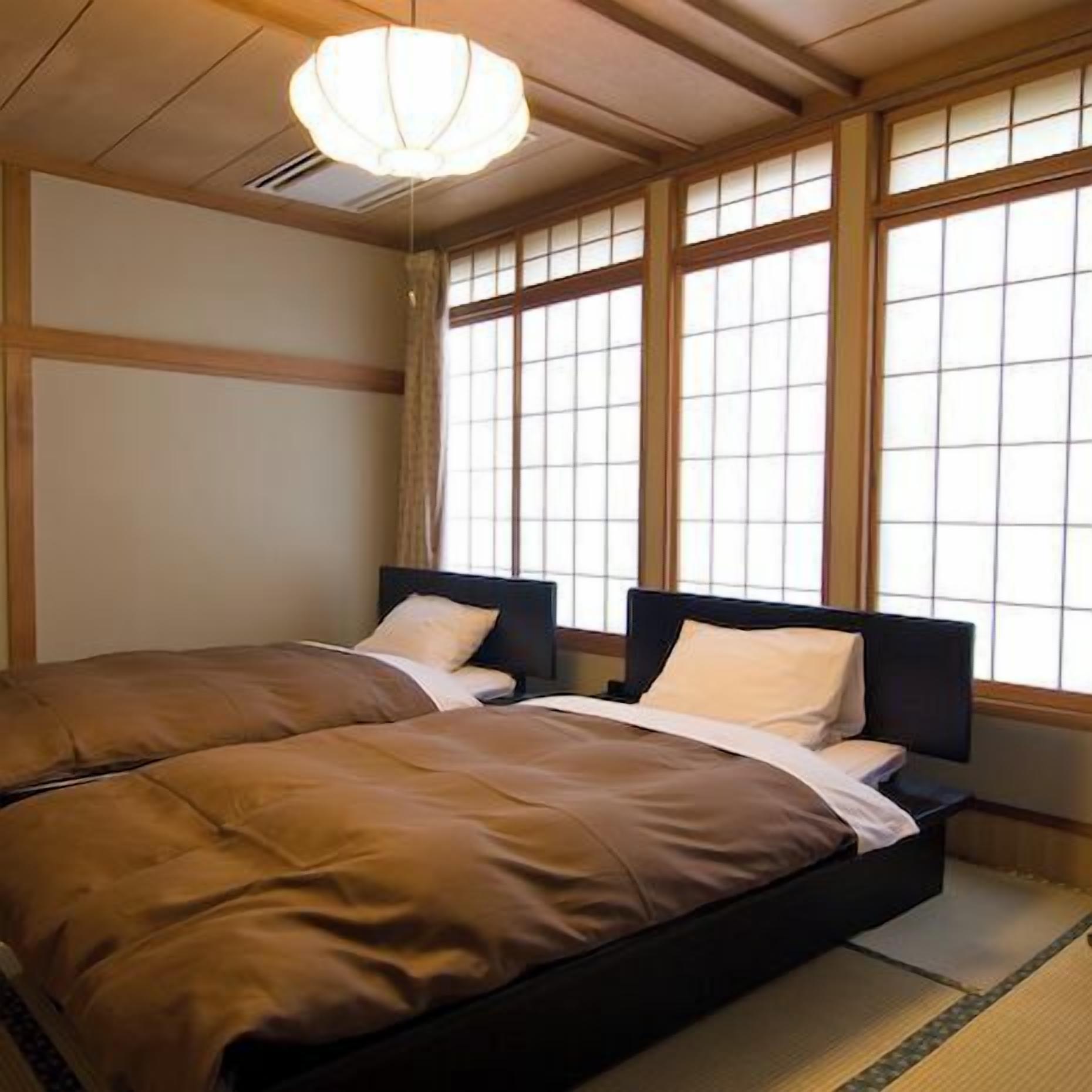 Una habitación tradicional japonesa con suelo de tatami, dos camas bajas con cabeceros oscuros y un ventanal con pantalla shoji.