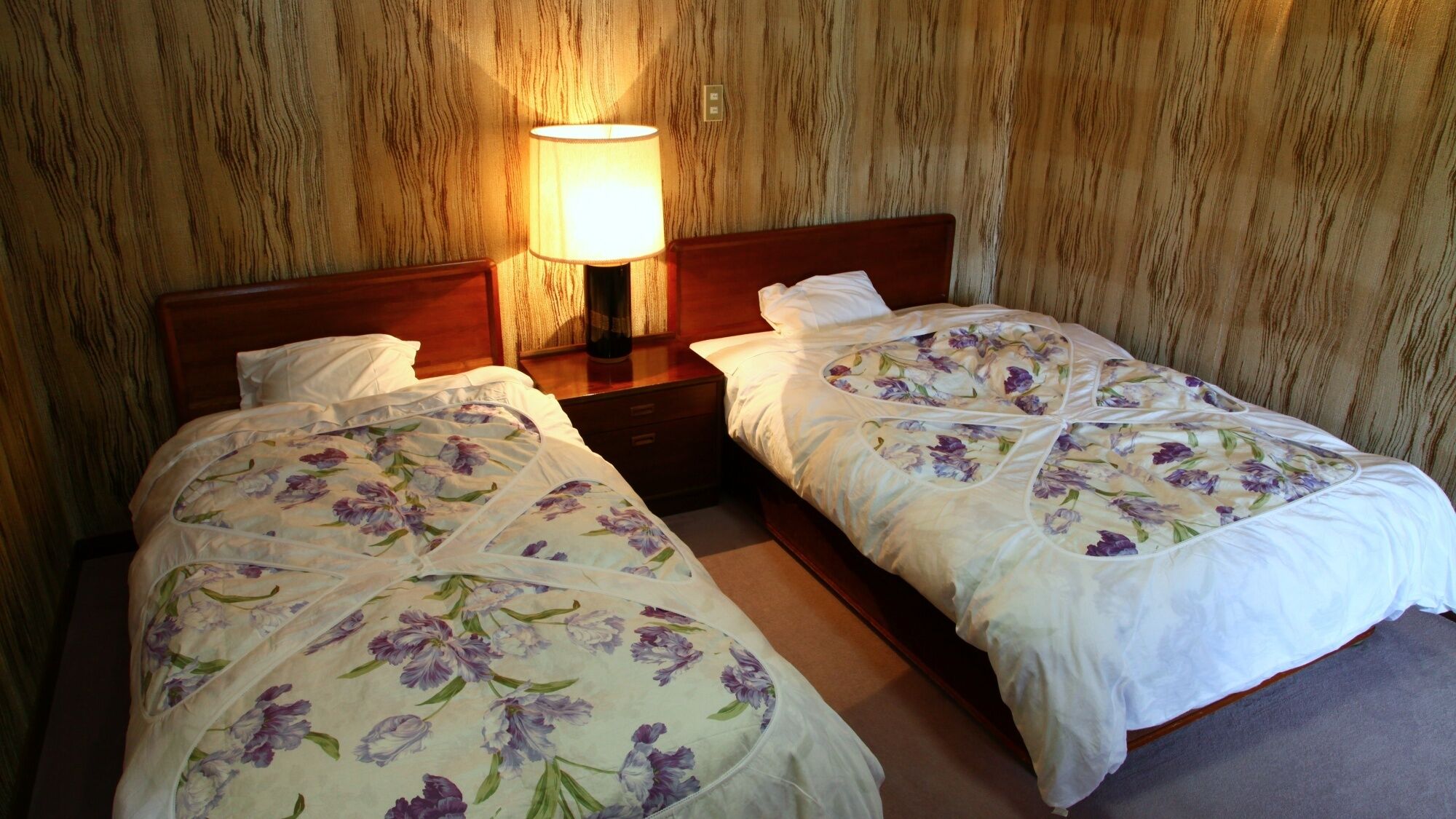 Two single beds with floral bedding, a nightstand with a lamp, and a wooden wall.