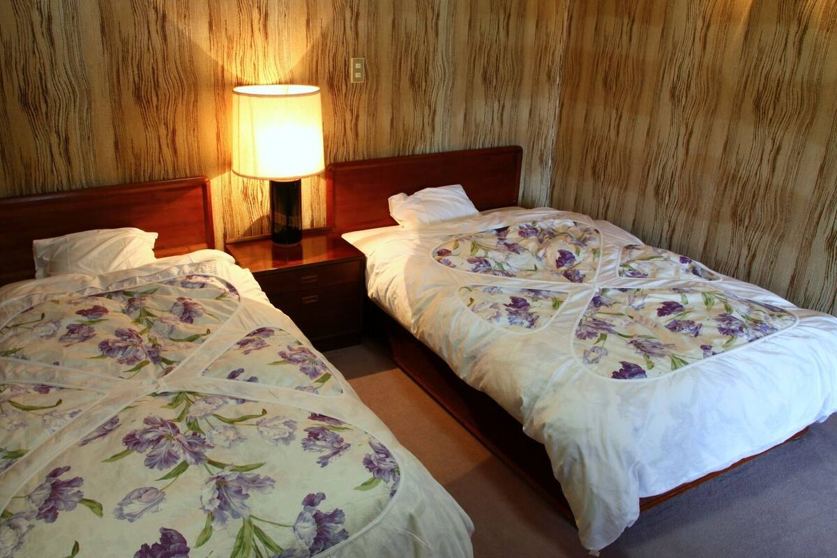 Two single beds with floral bedding, a nightstand with a lamp, and a wooden wall.