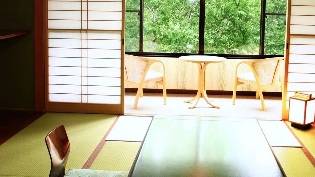 A tatami mat room with a low table and two chairs, a window with a view of trees, and a small shelf with books.