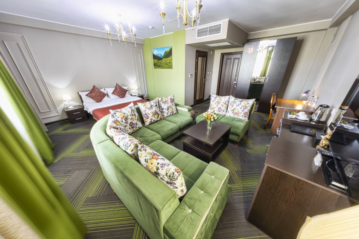 A modern hotel room with a green sectional sofa, a desk with a computer, and a bed with a red blanket.