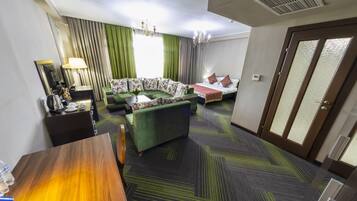 Habitación de hotel con sofá verde, cama con almohadones estampados, mesa de madera, espejo y candelabro.