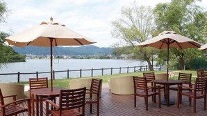 An outdoor deck with wooden tables and chairs, surrounded by umbrellas, overlooking a lake with a distant mountain view. - Lake Side Hotel & Restaurant Little White Flower (Kyotango)