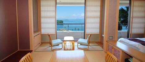 A hotel room with a wooden table, two chairs, a bed, and a view of the sea through large windows.