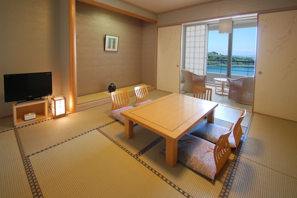 Une pièce de style japonais traditionnel, avec un sol recouvert de tatamis, une table basse, des chaises, un téléviseur et une vue sur la mer par une porte coulissante.